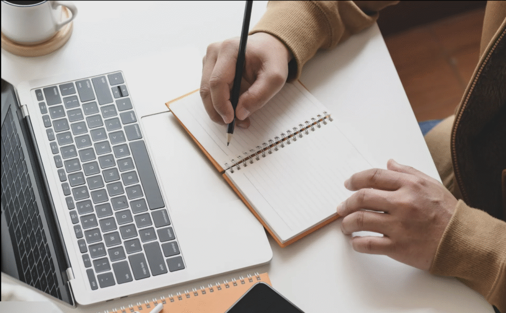 A laptop displaying a digital document with notes in progress next to a notepad, representing early-stage instructional design reflection.